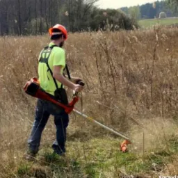 Mężczyzna w kasku i jaskrawej koszulce tnie trawę kosiarką spalinową w terenie zarośniętym suchymi trawami i chwastami, widoczne drobne odłamki trawy w powietrzu.