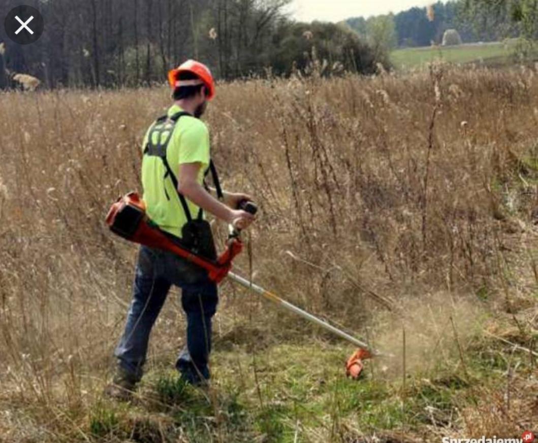 Mężczyzna w kasku i jaskrawej koszulce tnie trawę kosiarką spalinową w terenie zarośniętym suchymi trawami i chwastami, widoczne drobne odłamki trawy w powietrzu.