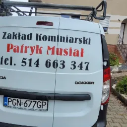 Tył białego samochodu Dacia Dokker z napisem 'Zakład Kominiarski Patryk Musiał tel. 514 663 347' na tle domu z ogrodem.