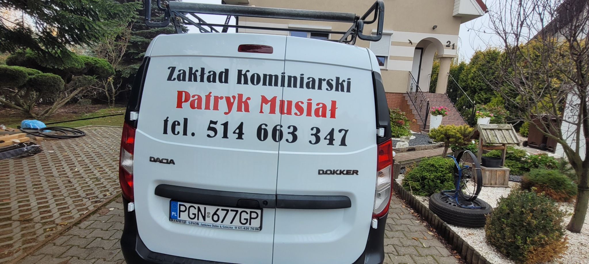 Tył białego samochodu Dacia Dokker z napisem 'Zakład Kominiarski Patryk Musiał tel. 514 663 347' na tle domu z ogrodem.