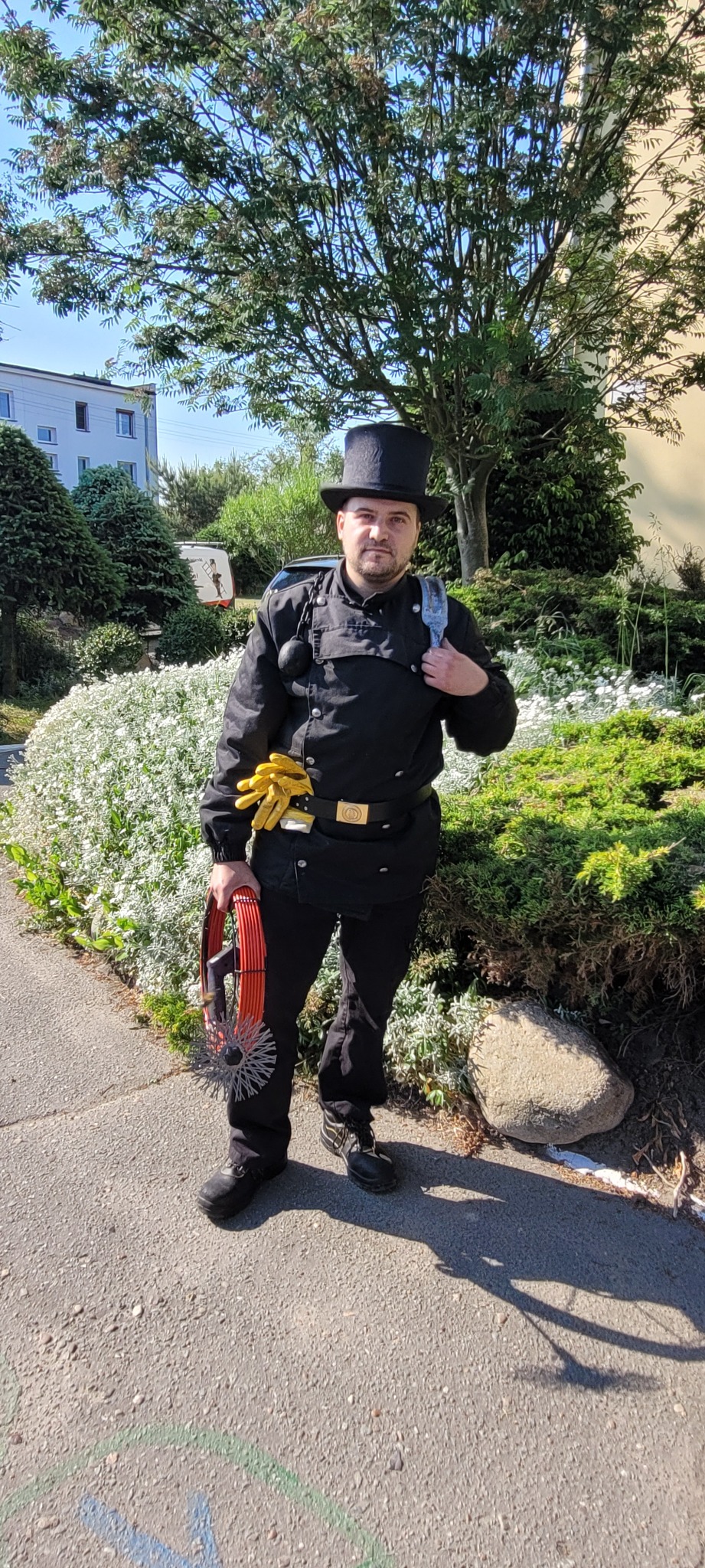 Uśmiechnięty kominiarz w pełnym stroju, z narzędziami w dłoni, pozuje na tle zieleni. Widoczny budynek w tle.