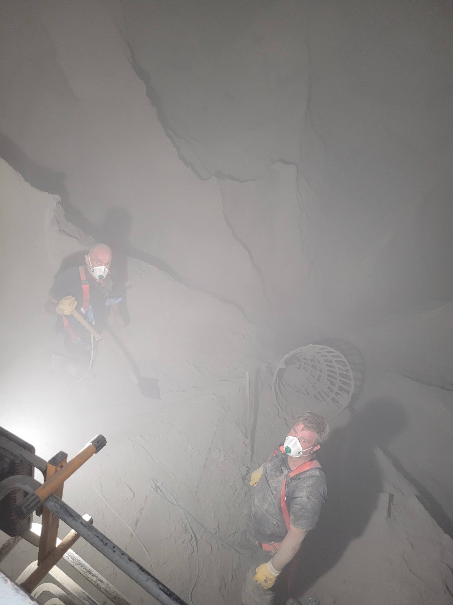 Dwóch kominiarzy w maskach i uprzężach, jeden z łopatą, pracuje w zadymionym kominie, widok z dołu na drabinę i wnętrze komina.