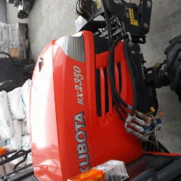Pomarańczowy ciągnik Kubota BX2350 z widocznymi przewodami hydraulicznymi i stertą worków w tle.