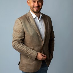 TOBIS Nieruchomości - Uśmiechnięty mężczyzna w brązowej marynarce i białej koszuli na jasnoszarym tle. Portret biznesowy z ręką w kieszeni, prezentujący profesjonalizm i pewność siebie.