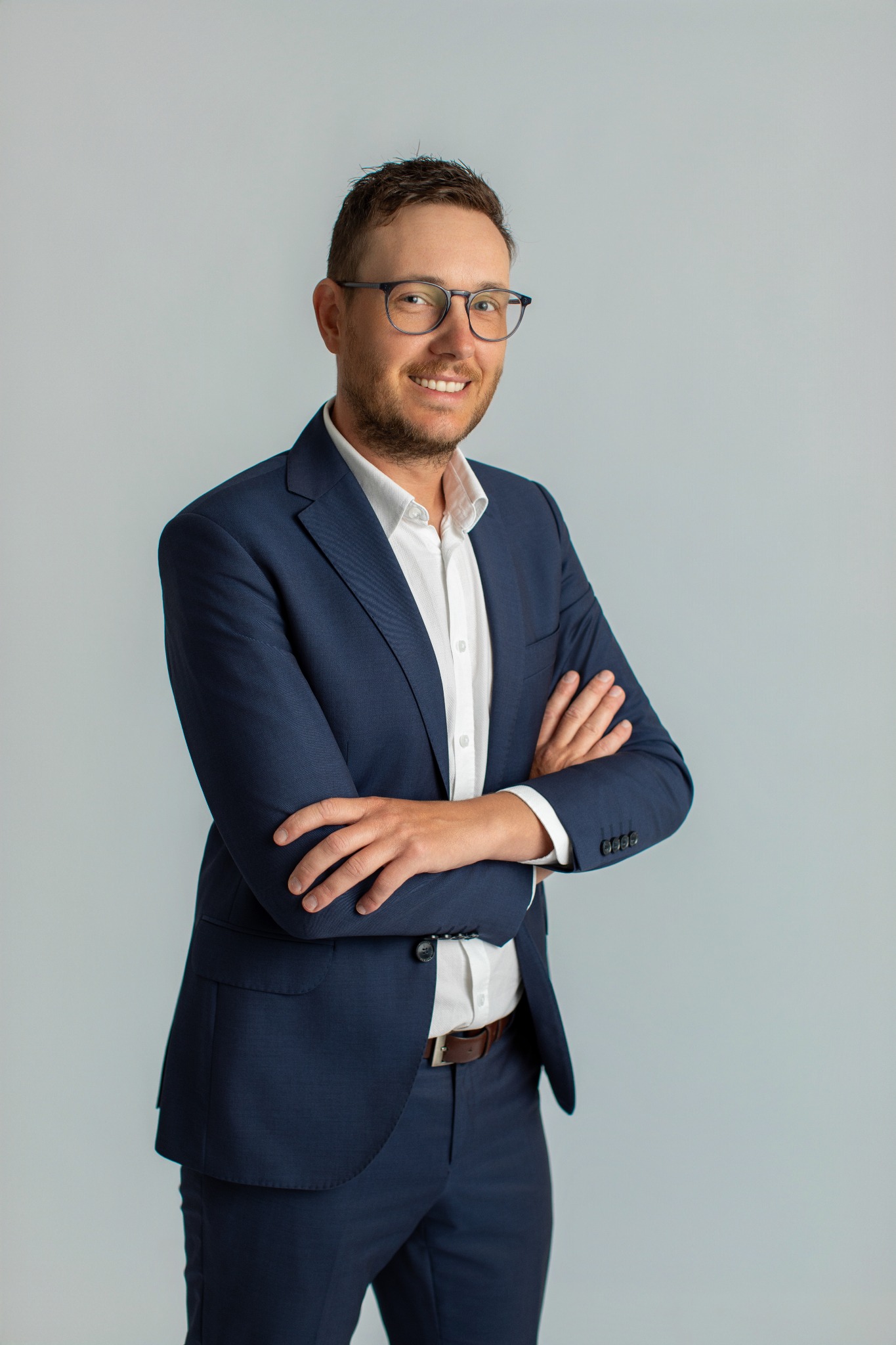 Uśmiechnięty doradca w okularach i granatowym garniturze, z założonymi rękami, prezentuje profesjonalizm i pewność siebie na neutralnym tle.
