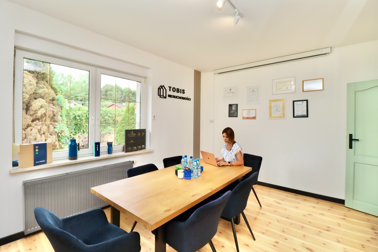 Wnętrze biura nieruchomości w Dąbrówce z kobietą pracującą na laptopie przy stole konferencyjnym, widok na zieleń za oknem, certyfikaty na ścianie.