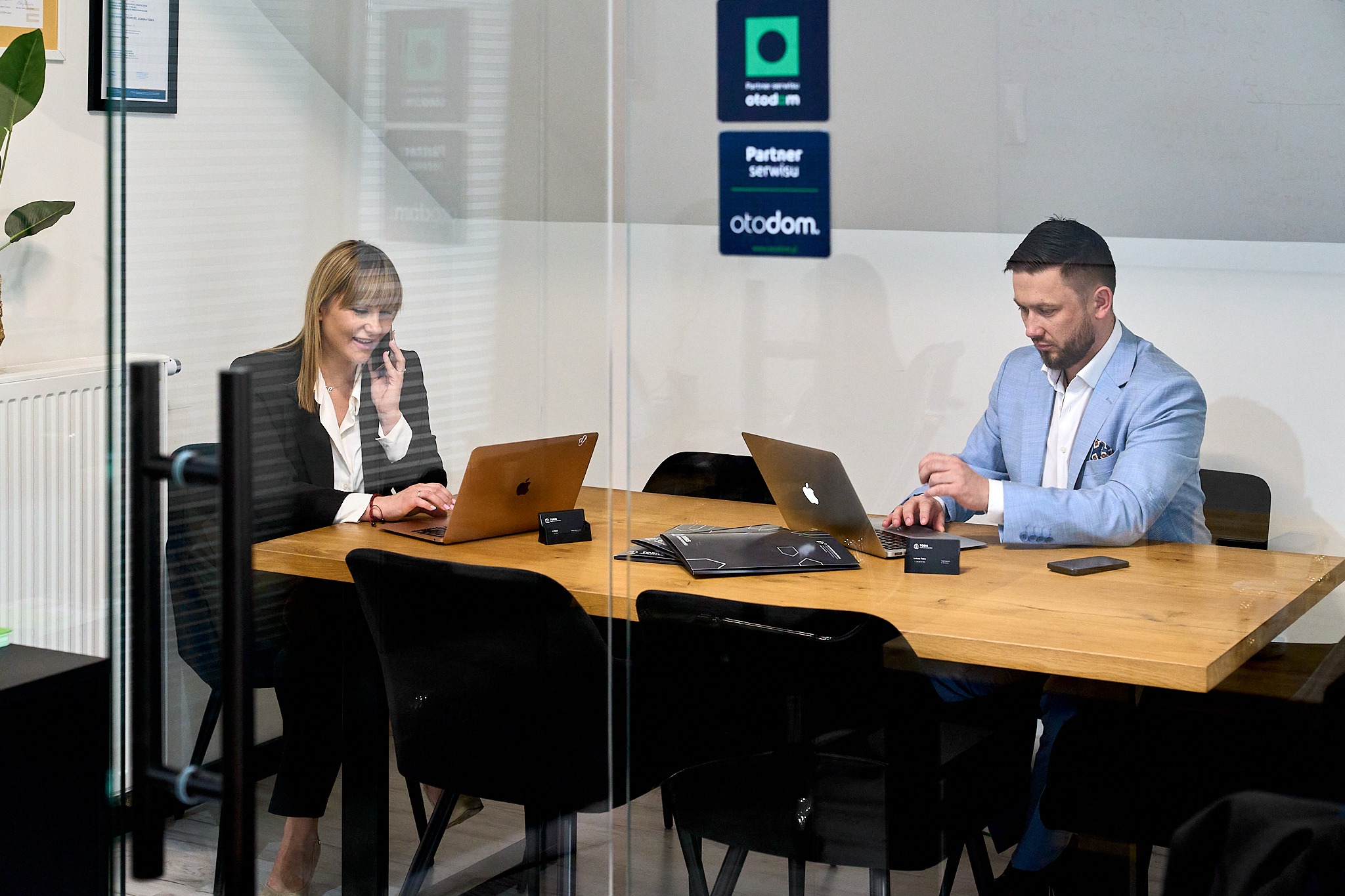 Dwoje konsultantów, kobieta rozmawia przez telefon, mężczyzna pracuje na laptopie przy biurku w nowoczesnym biurze z logo partnera serwisu Otodom na szklanej ściance.