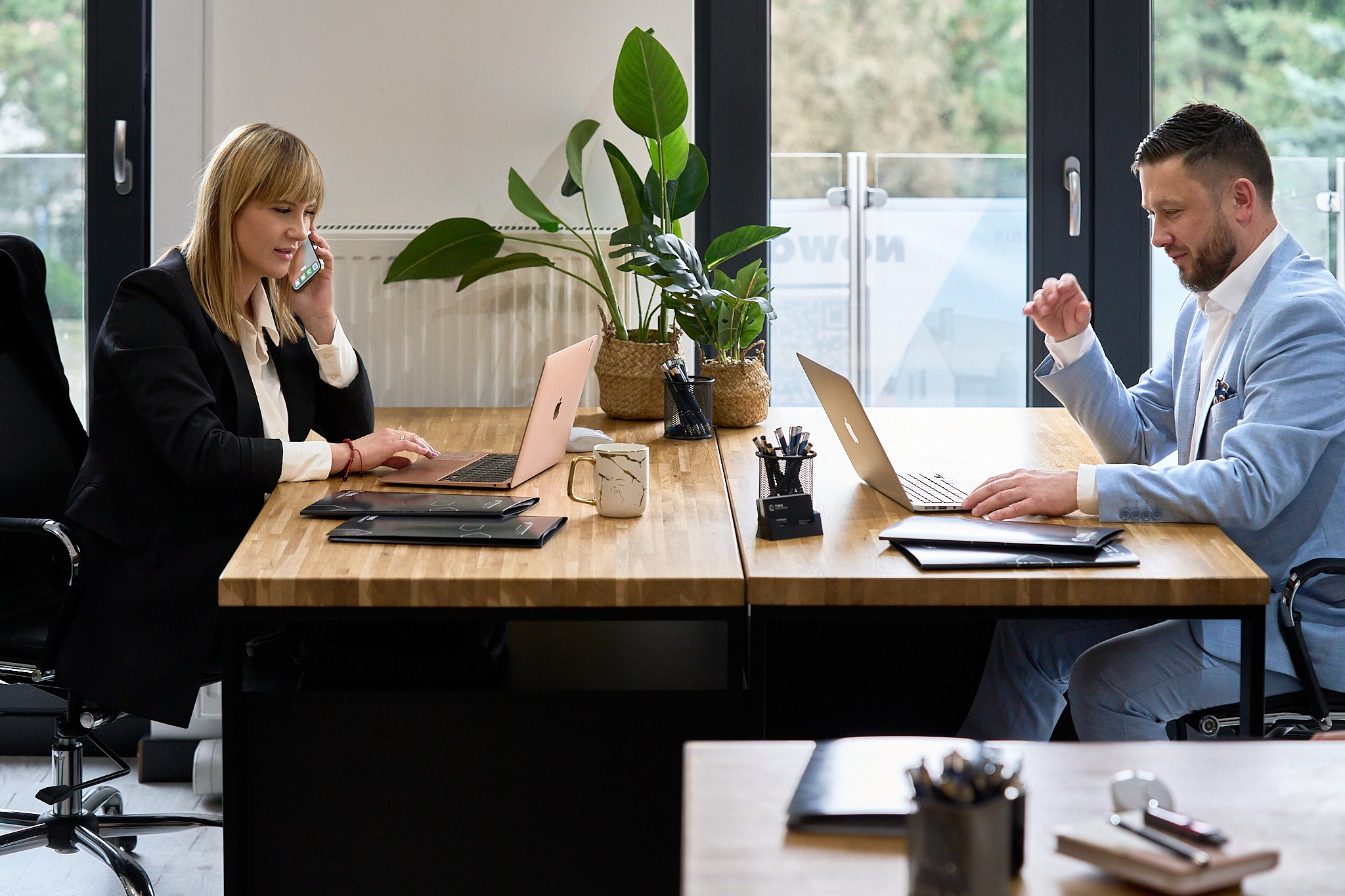Profesjonalne biuro: kobieta w trakcie rozmowy telefonicznej i mężczyzna pracujący na laptopie, na drewnianym biurku, z roślinami w tle.