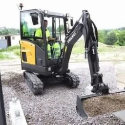 Kompaktowa koparka Volvo z operatorem w kabinie, łyżka wypełniona ziemią, widok z wnętrza budynku w trakcie budowy.