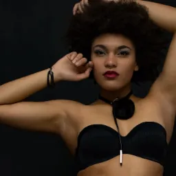 Portret kobiety z afro, w czarnym gorsecie i chokerze z pomponem na tle ciemnego studia, z wyrazistym makijażem oczu i ust.