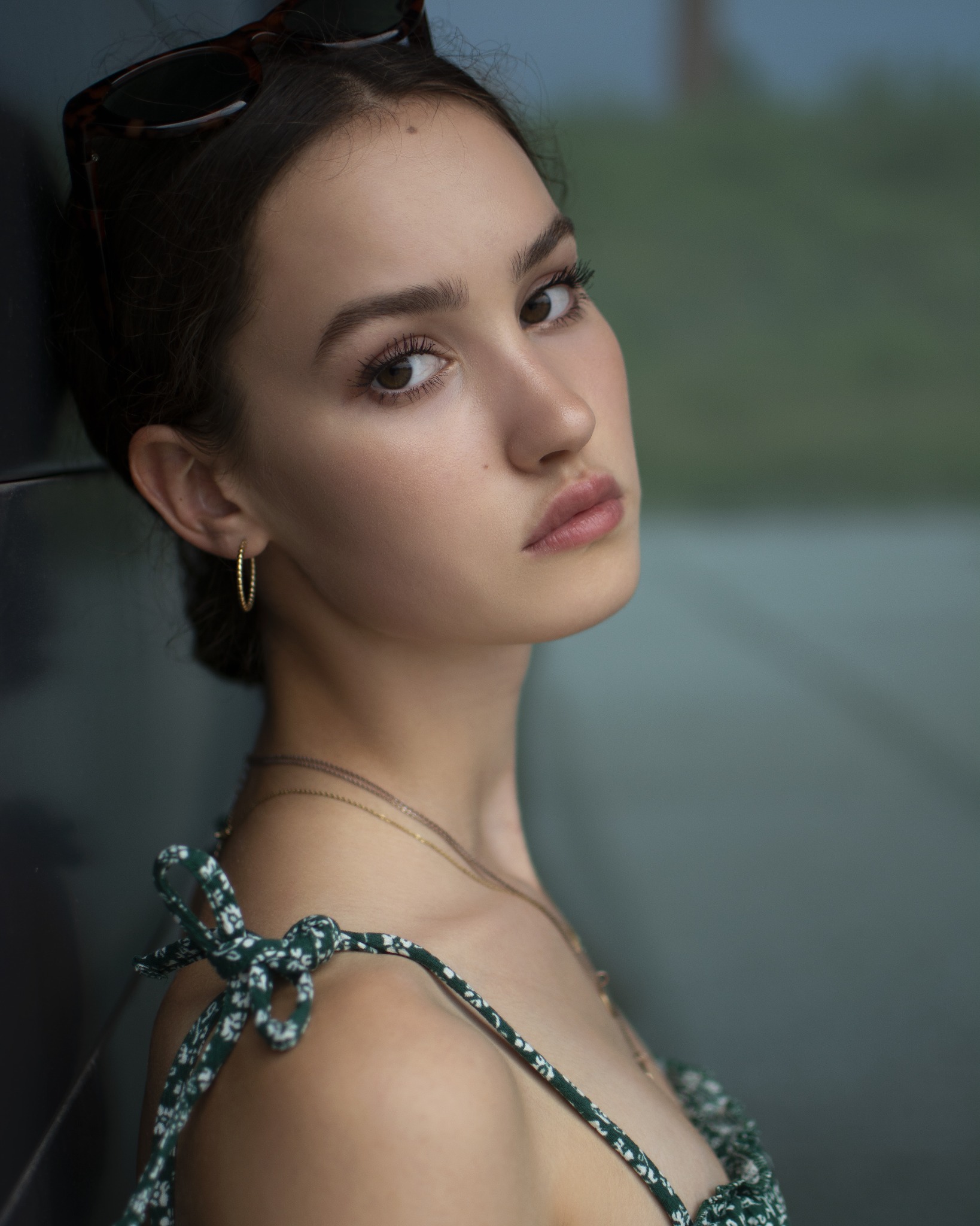 Portret młodej kobiety opierającej się o szybę, z okularami na głowie i delikatnym makijażem, ubrana w sukienkę na ramiączkach w drobne kwiatki, z widocznymi złotymi kolczykami i łańcuszkami