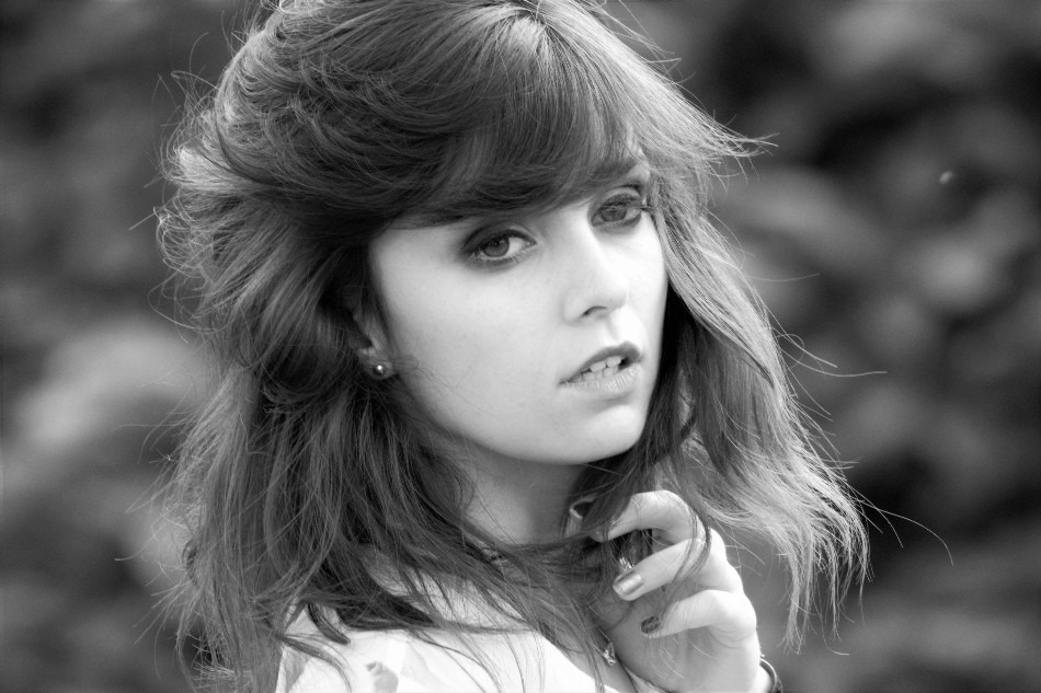 Portret czarno-biały młodej kobiety z rozwianymi włosami i delikatnym makijażem, spoglądającej w obiektyw z lekko otwartymi ustami, na rozmytym tle.