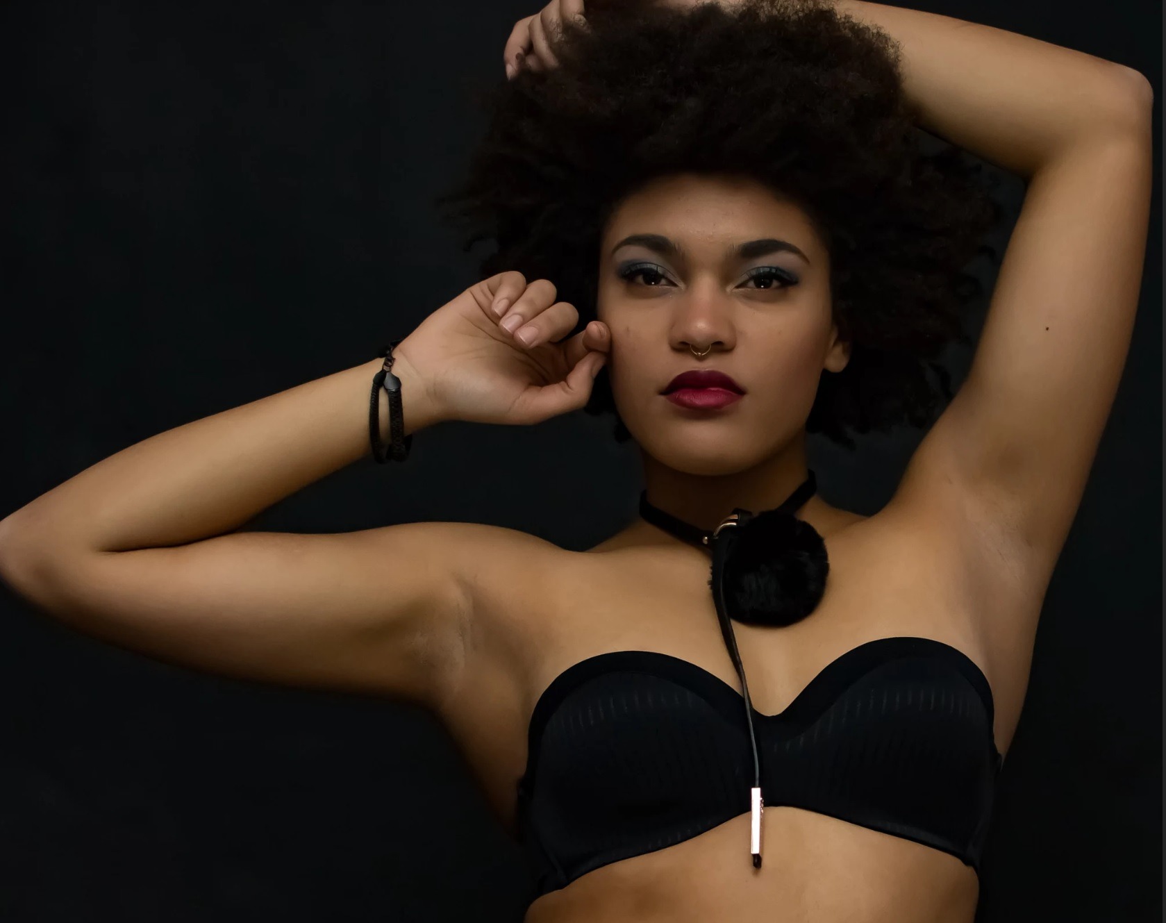 Portret kobiety z afro, w czarnym gorsecie i chokerze z pomponem na tle ciemnego studia, z wyrazistym makijażem oczu i ust.