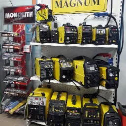 Ekspozycja różnych modeli spawarek MAGNUM i MONOLITH na półkach w sklepie, widoczne przewody i panele sterowania.