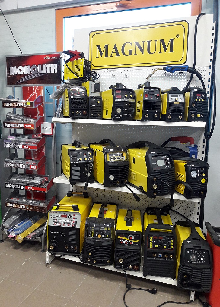 Ekspozycja różnych modeli spawarek MAGNUM i MONOLITH na półkach w sklepie, widoczne przewody i panele sterowania.