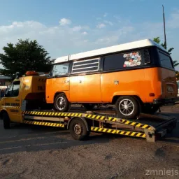 Żółta laweta z pomarańczowo-czarnym, oklejonym naklejkami vanem na platformie, widok z boku, w tle budynki.