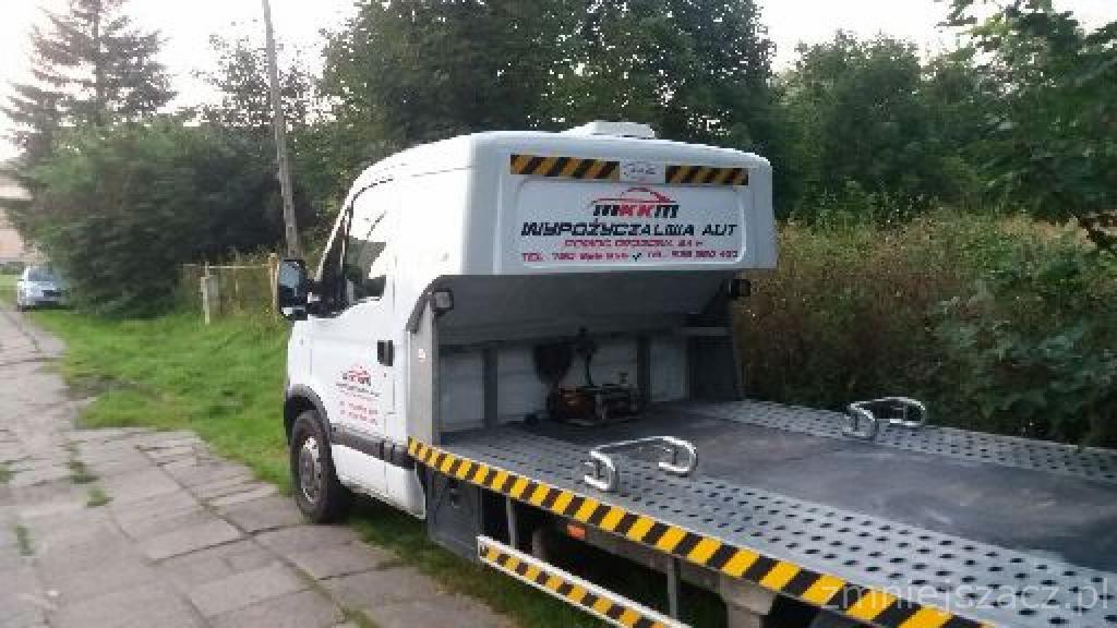 Biała laweta z żółto-czarnymi pasami ostrzegawczymi, widoczny pusty pokład i logo firmy 'Wypożyczalnia Aut' na nadbudówce