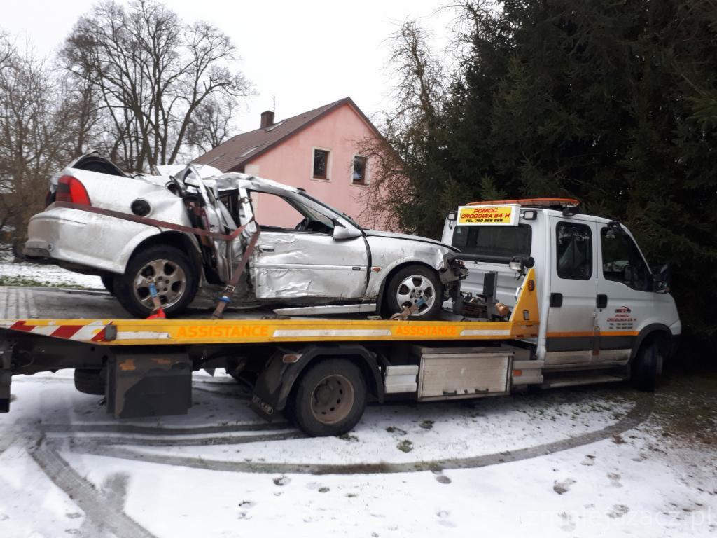 Laweta z uszkodzonym, srebrnym samochodem osobowym po wypadku, zabezpieczonym pasami transportowymi. Na lawecie widoczny napis 'ASSISTANCE'. W tle budynek mieszkalny i drzewa.