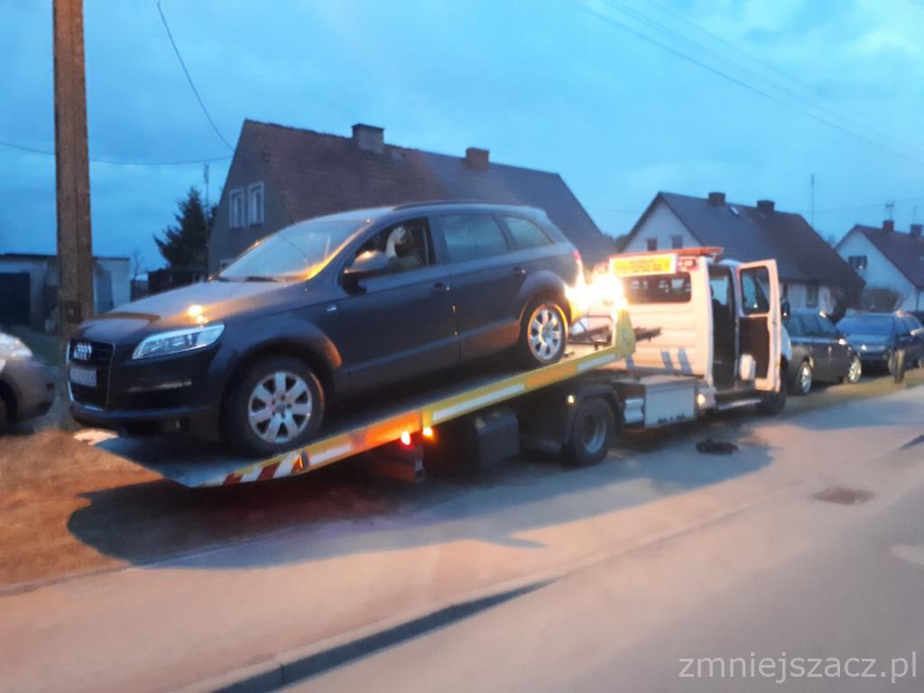 Granatowy SUV Audi Q7 na platformie lawety, widok z boku, światła lawety włączone, w tle zabudowa jednorodzinna.