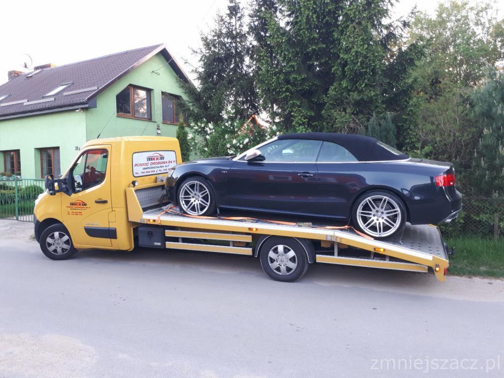 Żółta laweta z zaparkowanym na platformie ciemnym kabrioletem Audi A5 na tle zielonego domu i drzew.