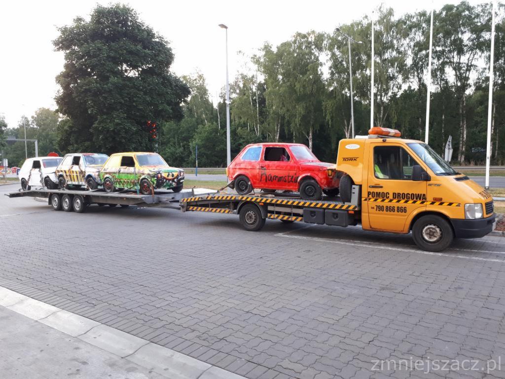 Żółta laweta z napisem 'Pomoc Drogowa' przewozi cztery pomalowane Fiaty 126p, jeden z napisem 'Rammstein', na tle drzew i chodnika.