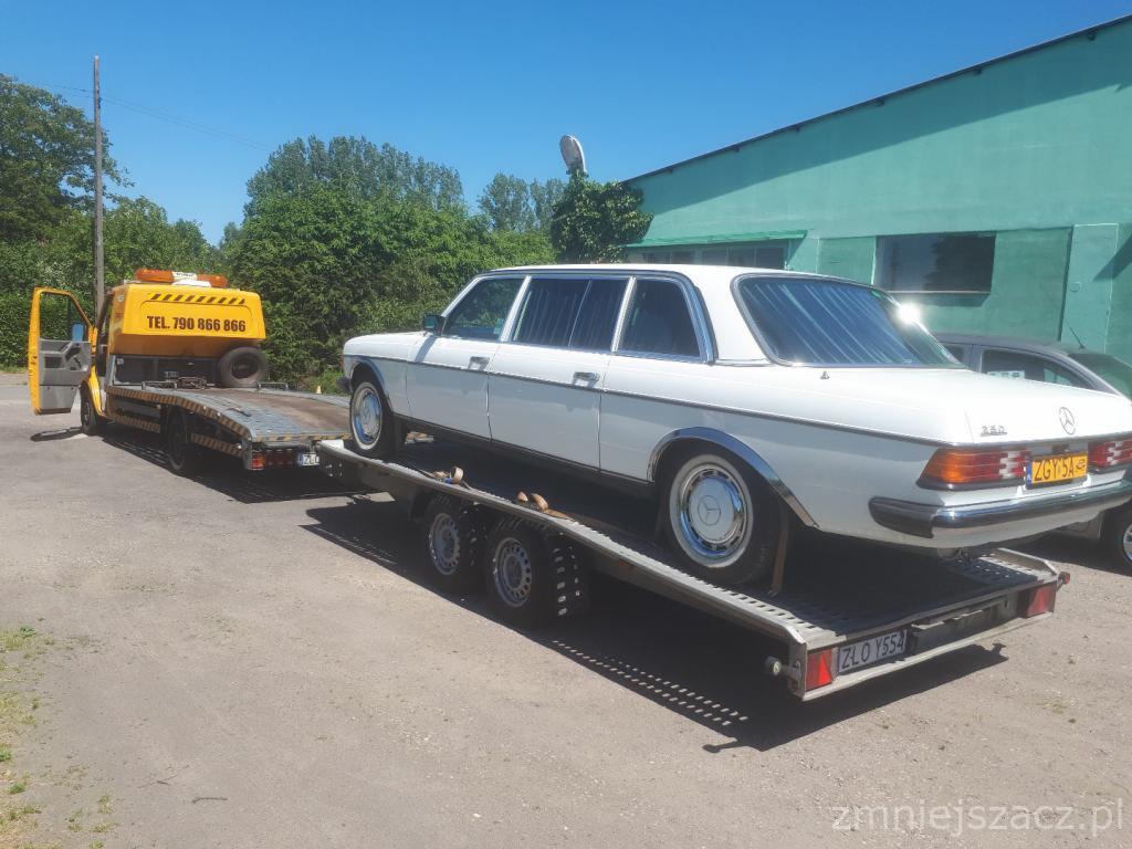 Żółta laweta z otwartą kabiną przewozi białego, zabytkowego Mercedesa W123 w wersji przedłużonej limousine na niskopodwoziowej przyczepie, widok z boku na tle zielonego budynku.