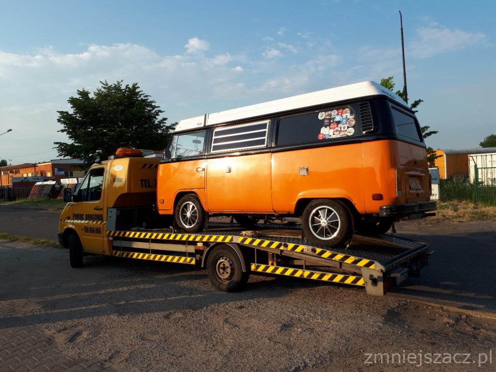 Żółta laweta z pomarańczowo-czarnym, oklejonym naklejkami vanem na platformie, widok z boku, w tle budynki.