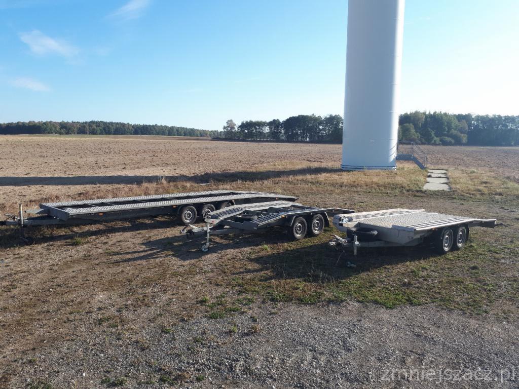 Trzy puste przyczepy lawety na polu w Resku, z wiatrakiem w tle, w słoneczny dzień.