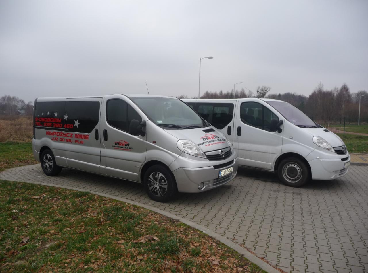 Dwa busy Opel Vivaro na parkingu; srebrny bus z naklejkami reklamowymi 'Wypożycz mnie już od 99 PLN' i logo firmy 'MKKM Wypożyczalnia Aut', drugi biały bus w tle.