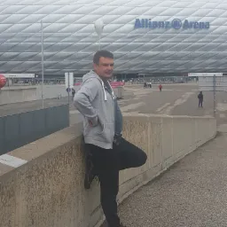 Mężczyzna w szarej bluzie z kapturem opiera się o betonową ścianę na tle stadionu Allianz Arena w Monachium.