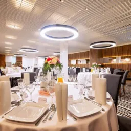 Uroczysta sala bankietowa z okrągłymi stołami nakrytymi białymi obrusami, elegancka zastawa, centralnie umieszczone kompozycje kwiatowe, nowoczesne okrągłe lampy sufitowe, ściany wykończone drewnem.