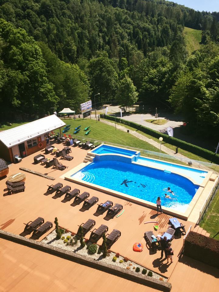 Widok z góry na basen hotelowy z błękitną wodą, otoczony leżakami i zielenią, w tle zalesione wzgórza.