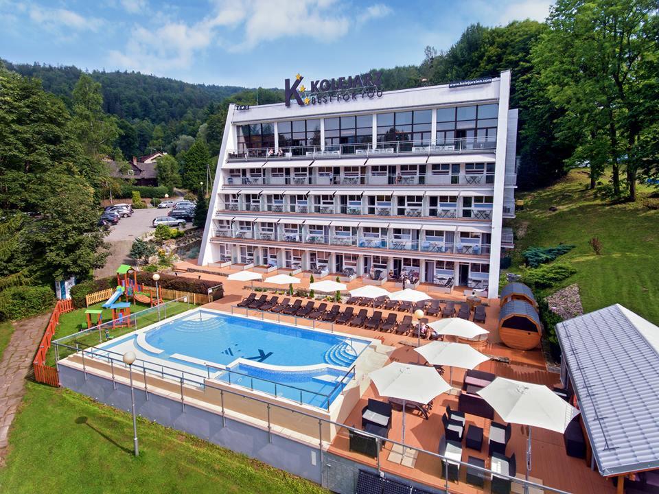 Wysoki budynek hotelowy z balkonami i basenem z leżakami, widok z góry na otoczenie zalesione.