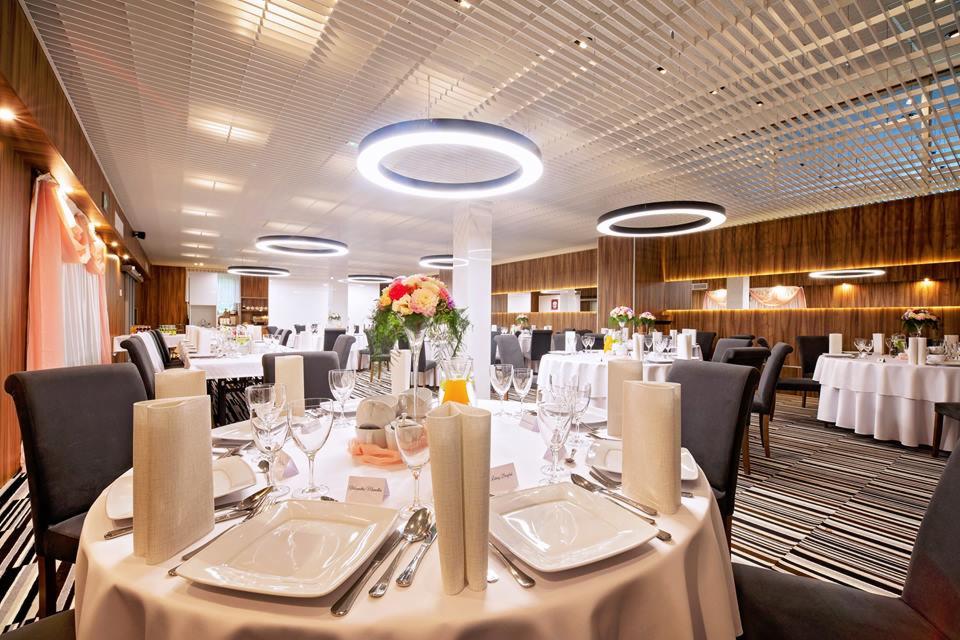 Uroczysta sala bankietowa z okrągłymi stołami nakrytymi białymi obrusami, elegancka zastawa, centralnie umieszczone kompozycje kwiatowe, nowoczesne okrągłe lampy sufitowe, ściany wykończone drewnem.