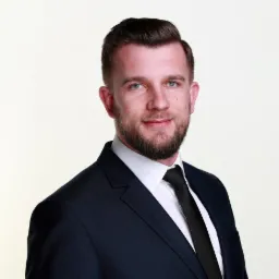 Portret młodego mężczyzny z brodą, ubranego w granatowy garnitur i czarny krawat, z delikatnym uśmiechem i jasnymi oczami na jasnym tle.