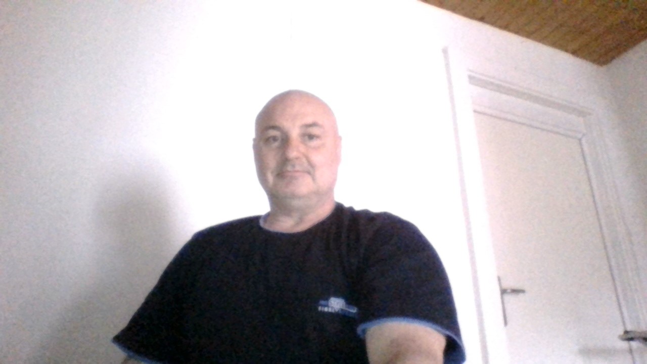 Portret mężczyzny na tle białej ściany i drzwi, z widocznym fragmentem sufitu z boazerią.