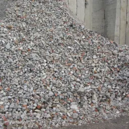 Góra gruzu z fragmentami cegieł i betonu na placu budowy, widoczne szare betonowe ściany w tle.