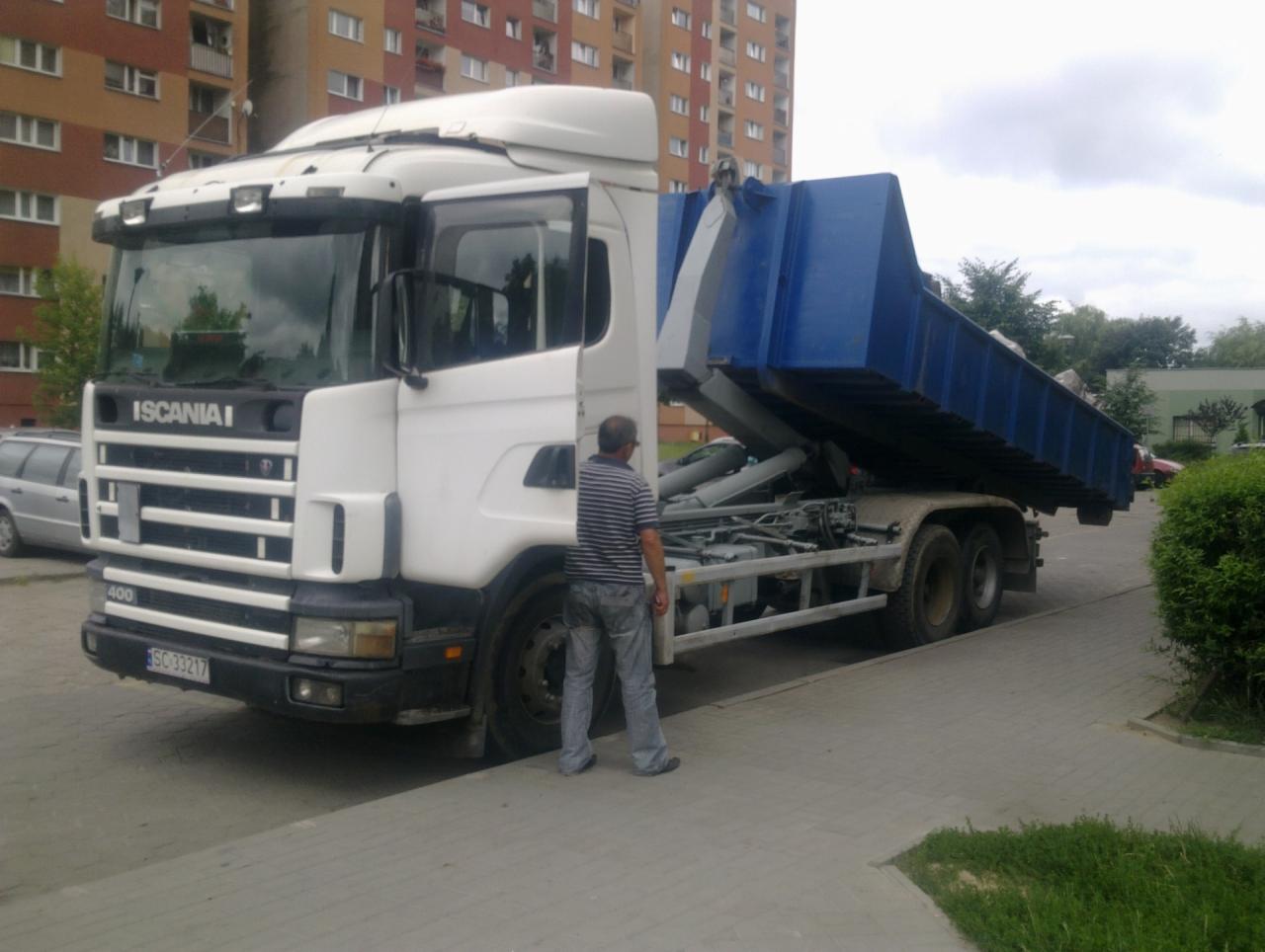 Biała ciężarówka Scania z podniesioną, niebieską skrzynią ładunkową na tle bloku mieszkalnego, widoczny mężczyzna w paski obserwujący proces.