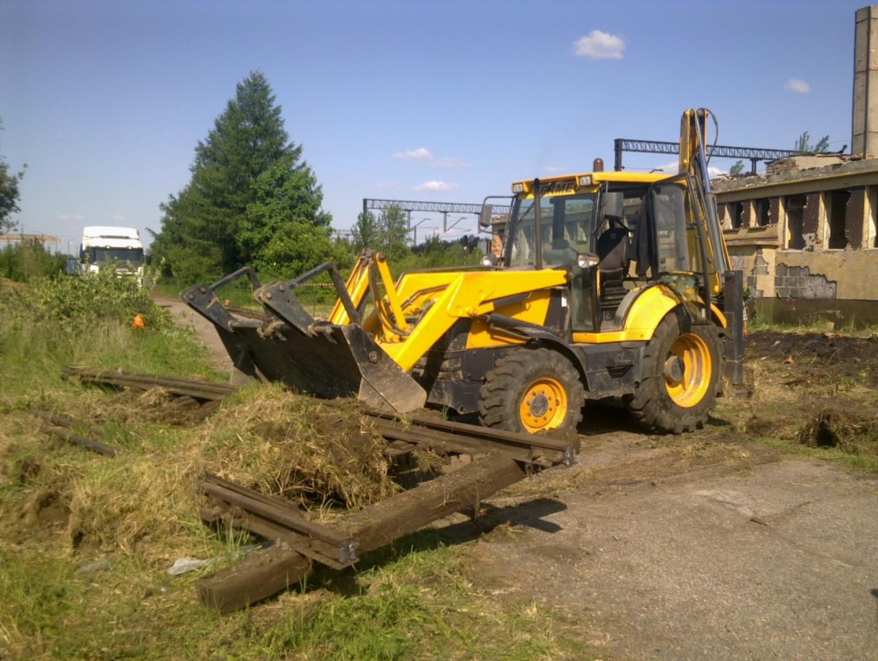 Żółta maszyna budowlana typu koparko-ładowarka zbiera resztki trawy i gruzu z metalowych szyn na terenie budowy z częściowo zrujnowanym budynkiem w tle.