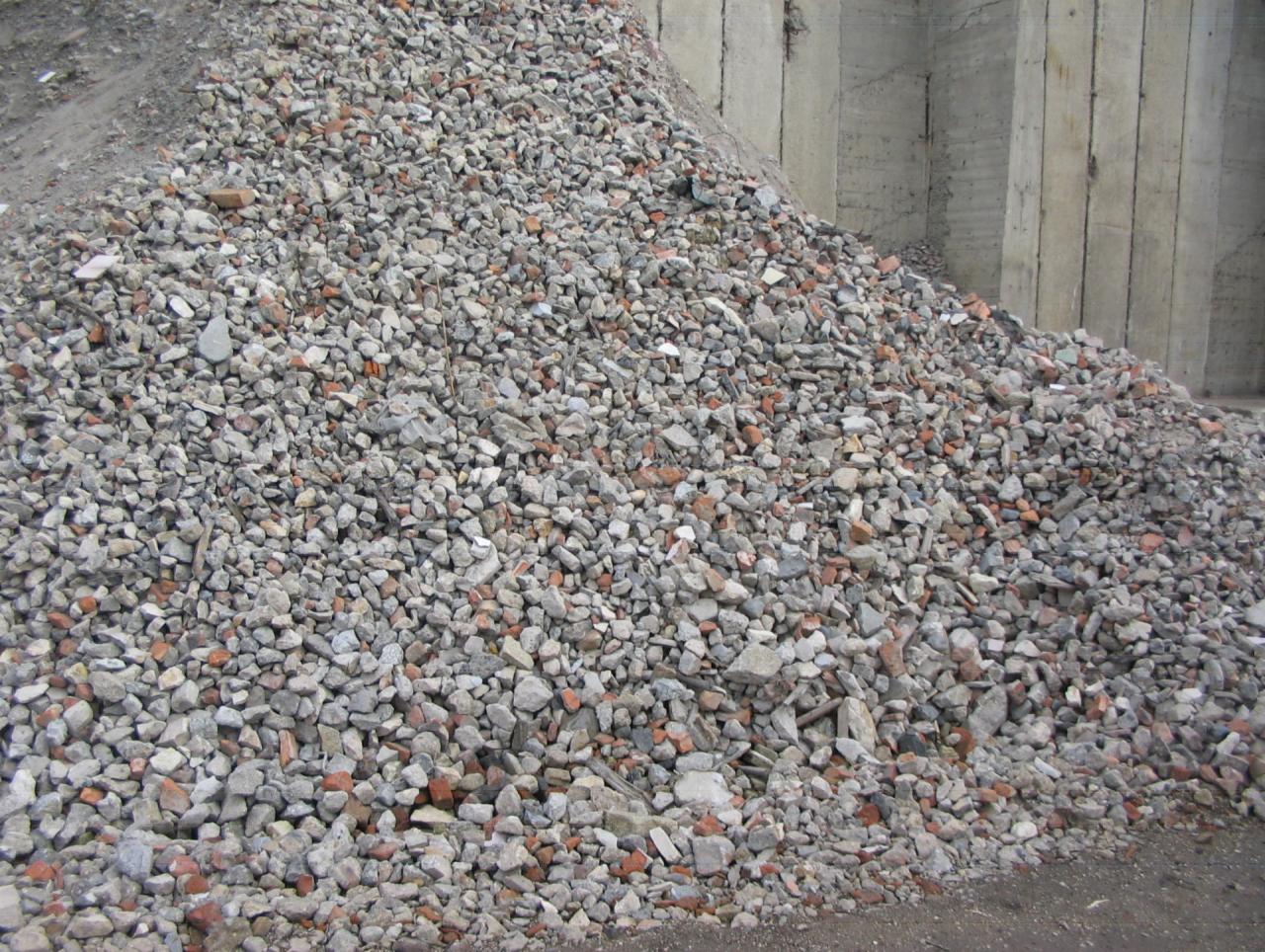 Góra gruzu z fragmentami cegieł i betonu na placu budowy, widoczne szare betonowe ściany w tle.