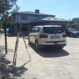 Pomiar geodezyjny przed domem: teodolit na trójnogu, biały samochód osobowy zaparkowany obok, kostka brukowa na podjeździe.