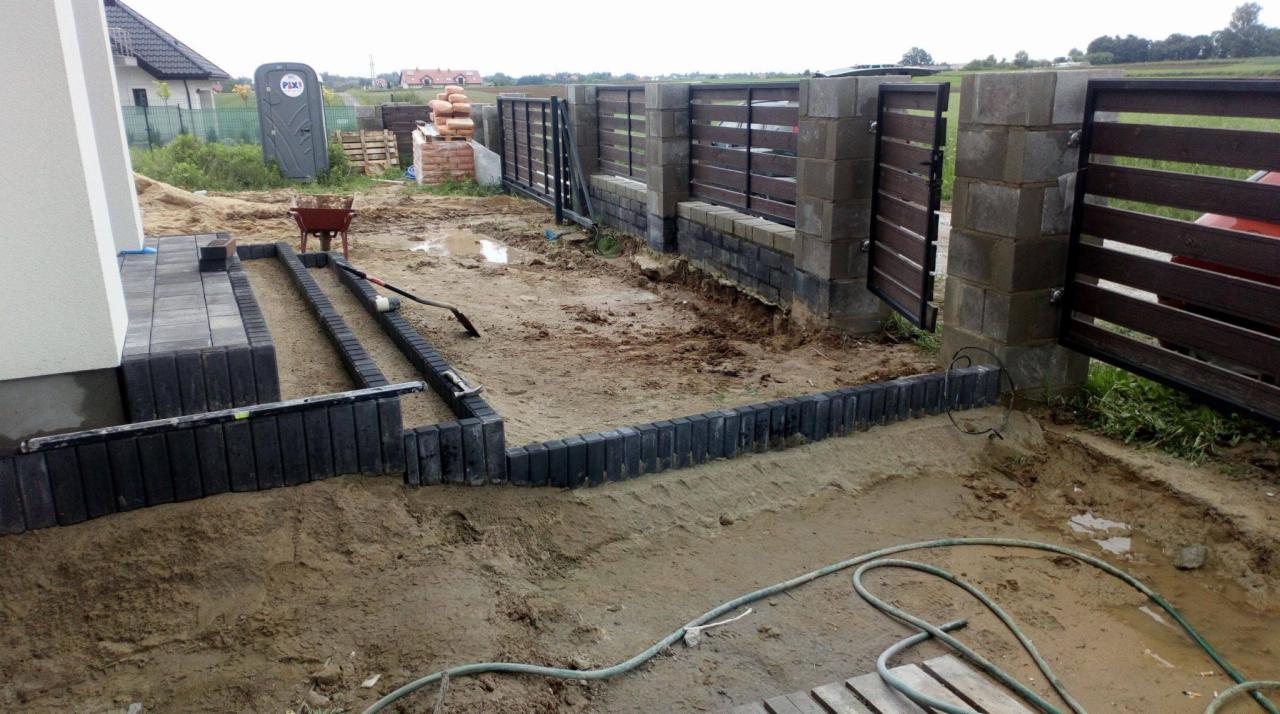 Układanie obrzeży z kostki brukowej na posesji, widoczne przygotowane podłoże, częściowo ułożona kostka przy domu, w tle ogrodzenie z drewnianymi przęsłami i przenośna toaleta.