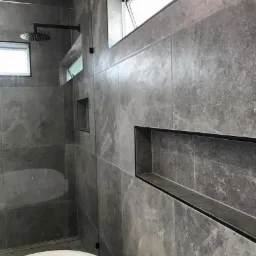 Nowoczesna łazienka z szarymi płytkami imitującymi kamień, czarną armaturą i wnęką w ścianie, widok na kabinę prysznicową z deszczownicą.