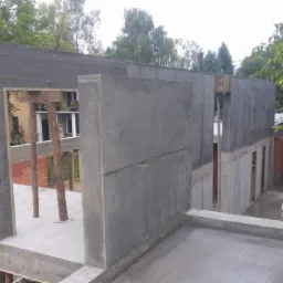 Surowy stan budynku w trakcie budowy z widocznymi betonowymi ścianami i drewnianymi podporami, ceglaną ścianką działową oraz drzewami w tle.