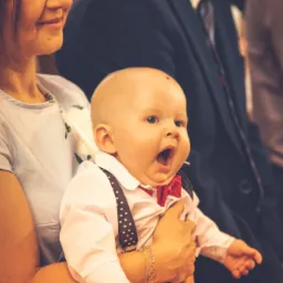 Uśmiechnięta kobieta trzyma na rękach małego chłopca w białej koszuli, czerwonej muszce i szelkach w kropki, kt&oacute;ry szeroko otwiera buzię, w tle mężczyzna w garniturze