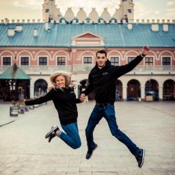 M-Foto Usługi Fotograficzne - Uśmiechnięta para w czarnych bluzach i jeansach skacze trzymając się za ręce na tle Zamku Lubelskiego, oboje w ruchu, dynamiczne ujęcie.