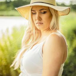 M-Foto Usługi Fotograficzne - Portret kobiety w ciąży w białej sukience i słomkowym kapeluszu na tle zieleni i jeziora, z delikatnym oświetleniem i subtelnym wyrazem twarzy.