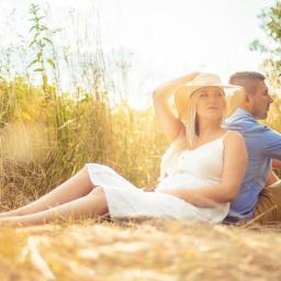 M-Foto Usługi Fotograficzne - Ciężarna kobieta w białej sukience i kapeluszu siedzi w polu, opierając się plecami o mężczyznę w błękitnej koszuli, ciepłe światło słoneczne.