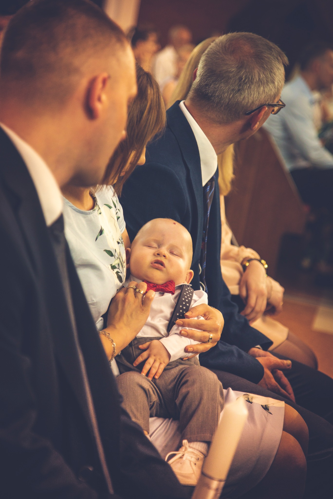Śpiące dziecko w stroju galowym z czerwoną muszką, trzymane na rękach podczas uroczystości w kościele, widoczni goście w tle.