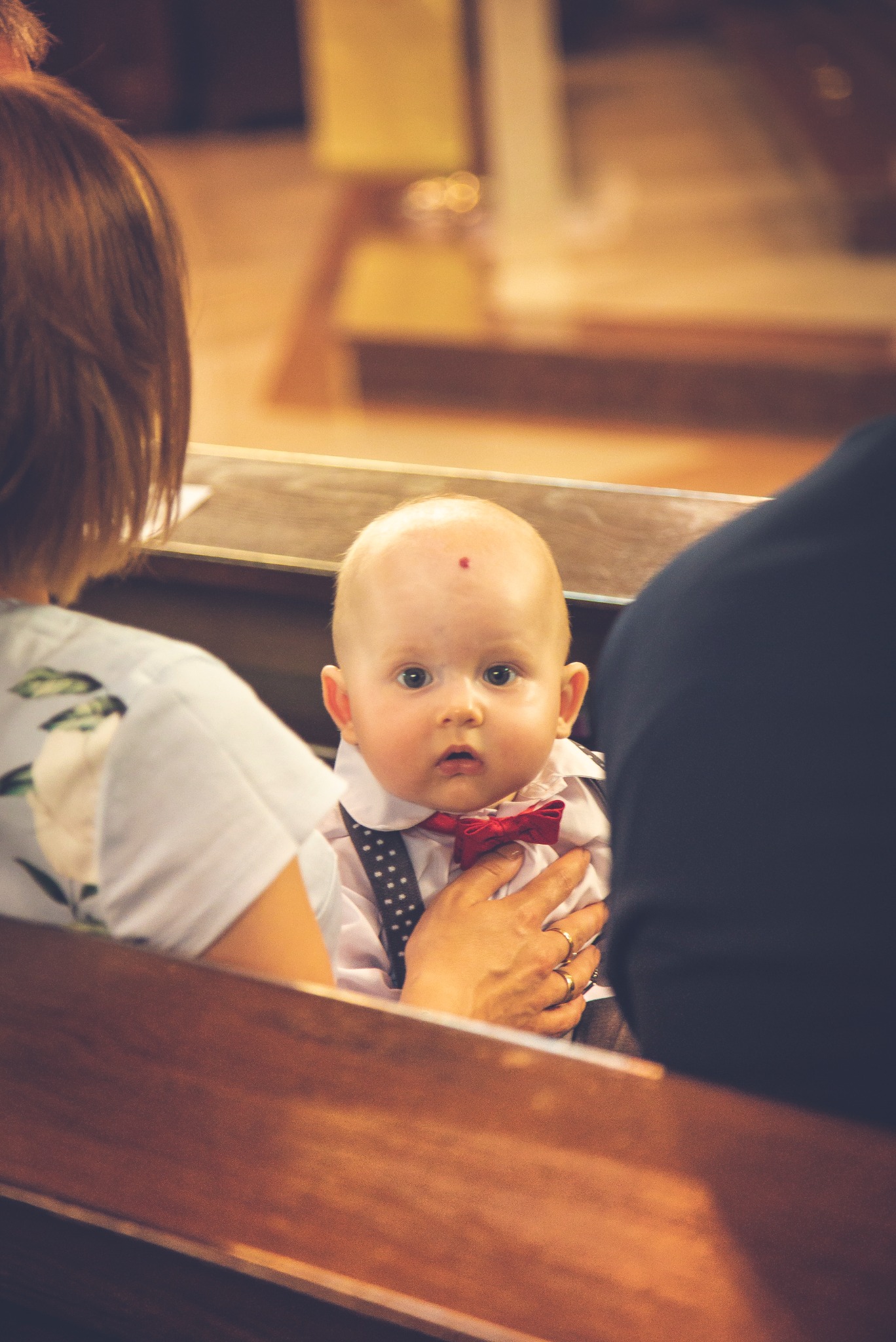 Uroczy niemowlak w eleganckim stroju z muchą, trzymany na rękach podczas uroczystości w kościele, z widocznym znakiem na czole.